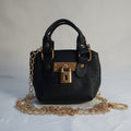 A small black pebbled leather handbag with gold-tone hardware, featuring a top handle, a detachable gold chain strap, and a lock closure, laid on a white fabric background.