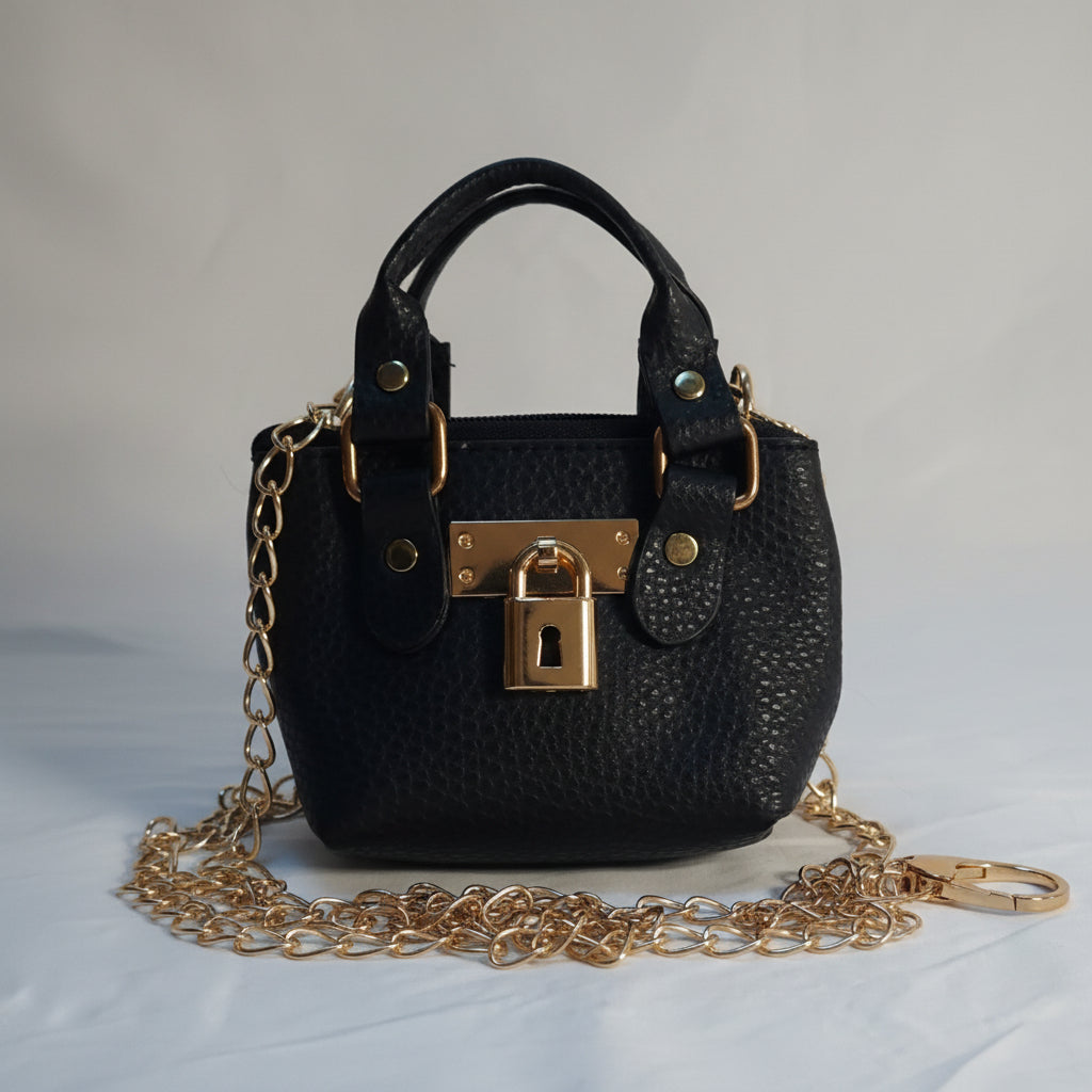 A small black pebbled leather handbag with gold-tone hardware, featuring a top handle, a detachable gold chain strap, and a lock closure, laid on a white fabric background.