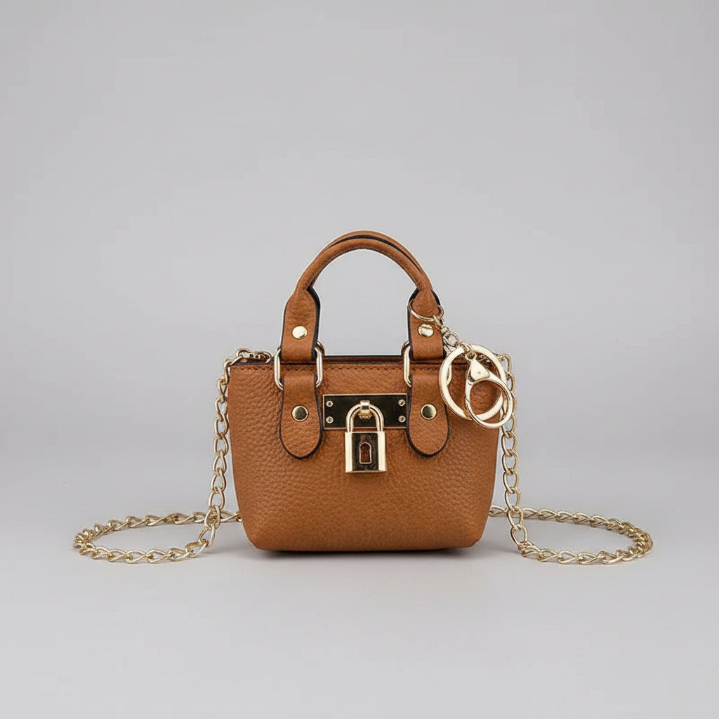 A small tan pebbled leather handbag with a gold chain strap, padlock detail, and double O-ring hardware, held by a hand with red nail polish against a white textured background.